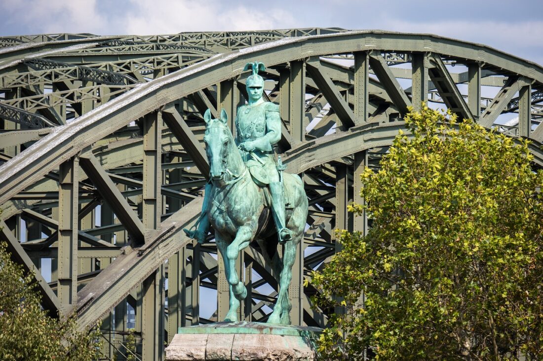 im Vordergrund ist ein Pferd mit Kaiser Wilhelm II als Reiterstandbild, dahinter zeigt sich der stählerne Bogen einer Rheinbrücke. Links neben dem Standbild sieht man einen blühenden Strauch