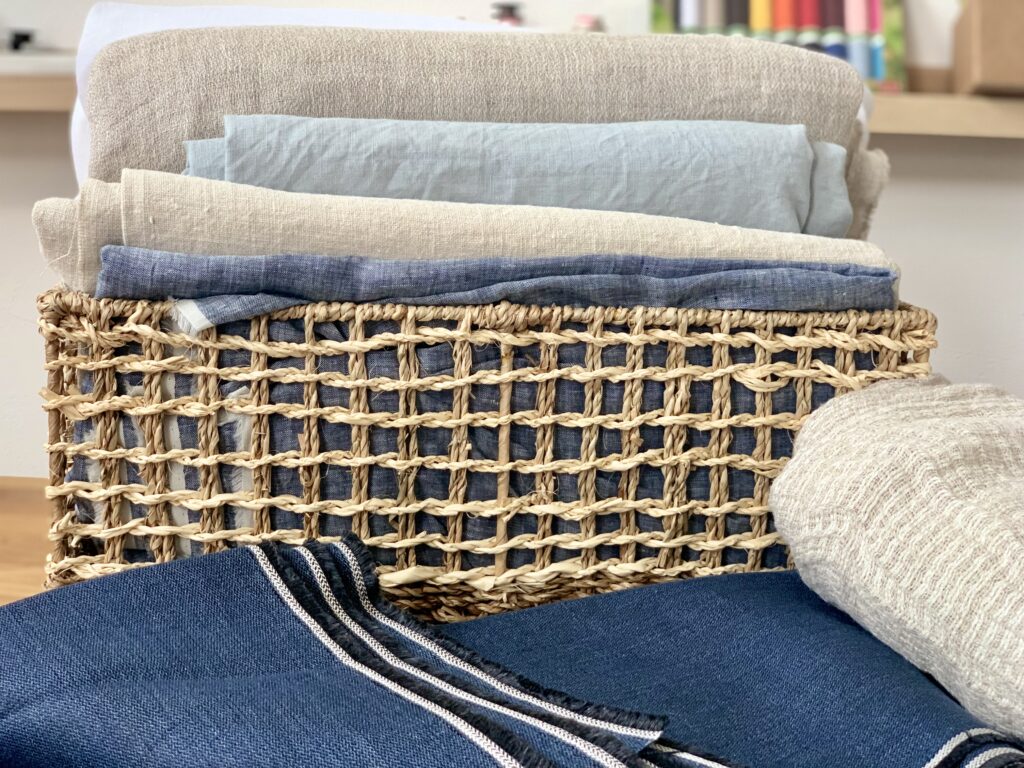 verschiedene Leinenstoffe in einem Korb, davor weitere Leinengewebe, im Hintergrund ist verschwommen ein kleines Regal zu sehen